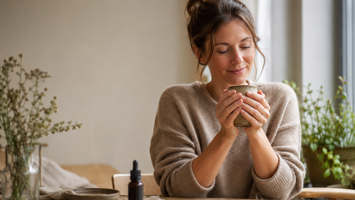 Frau sitzt am Fenster und wärmt ihre Hände an einer Keramiktasse, während eine CBD-Öl-Flasche unscharf auf dem Tisch steht – ruhige Wellness-Szene zum Thema CBD bei Rheuma.