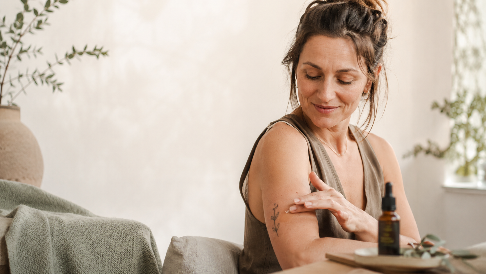 Frau trägt behutsam CBD-Öl auf ein kleines Tattoo am Oberarm auf – ruhige Tattoo-Nachpflege in heller, natürlicher Wellness-Atmosphäre.