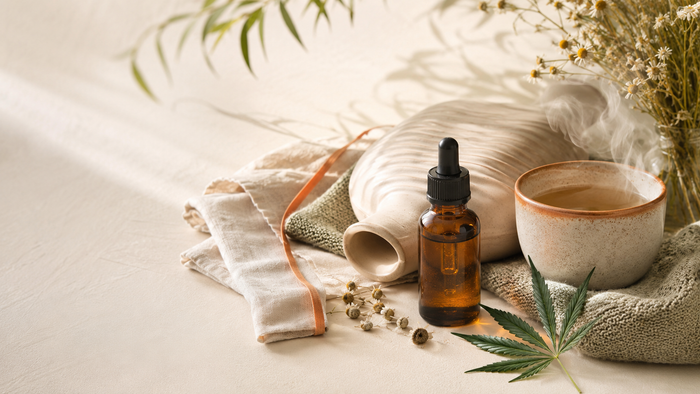 Amber CBD oil dropper bottle beside warm herbal tea, soft linen, ceramic hot water bottle, chamomile, and subtle hemp leaf in a calming wellness still life.