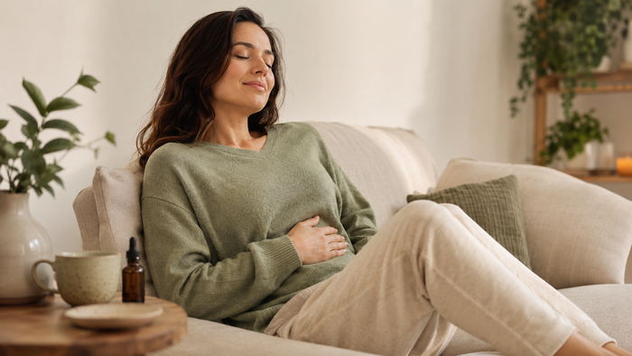 Frau entspannt sich auf einem Sofa mit einer Hand auf dem Bauch, daneben stehen CBD-Öl und Kräutertee in einer ruhigen, warmen Wohnatmosphäre.