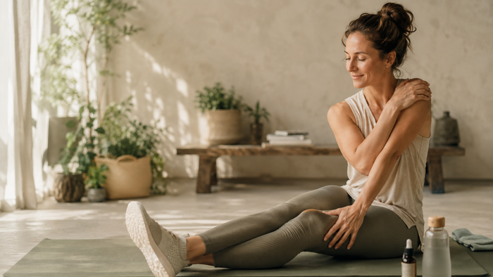 Frau entspannt sich nach dem Sport auf einer Yogamatte und dehnt ihre Schulter, während eine CBD-Öl-Flasche und eine Wasserflasche daneben liegen – natürliche Regeneration und Muskelentspannung.