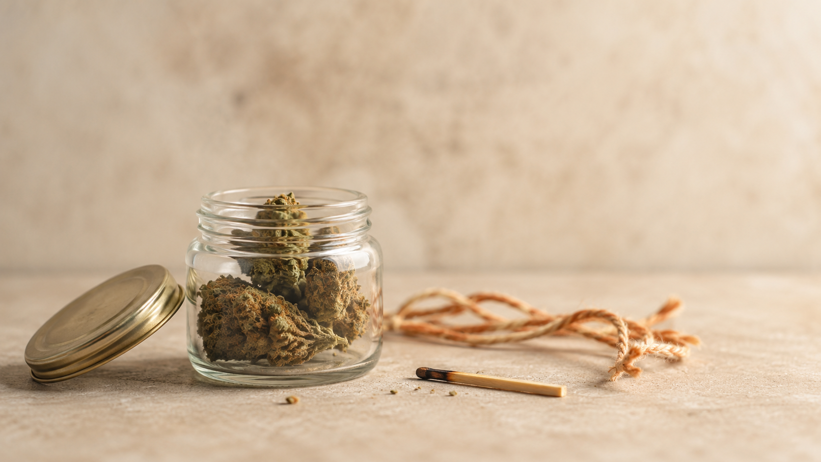 Offenes Glas mit getrockneten THCA-Blüten auf einer hellen Steinoberfläche neben einem einzelnen angekohlten Streichholz und einer Naturkordel – ruhige, sachliche Darstellung der Umwandlung von THCA zu THC durch Hitze.